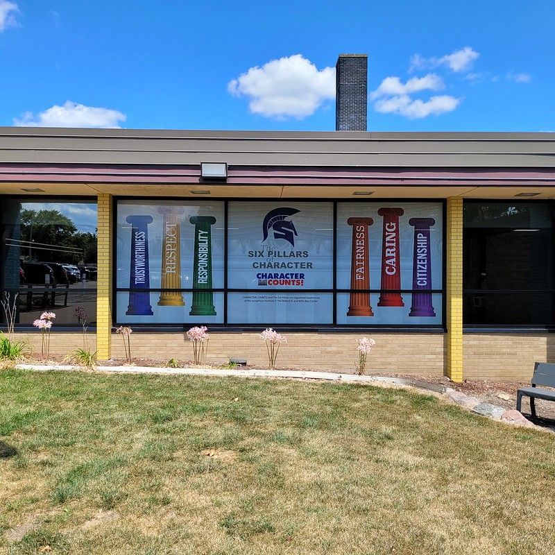 A school exterior featuring a large window decal displaying six colorful pillars labeled with character traits and a mascot.