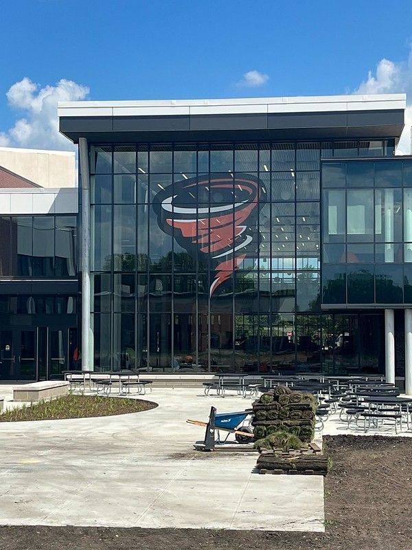Modern building exterior featuring a large, red tornado logo on a glass facade above a paved patio with outdoor tables.