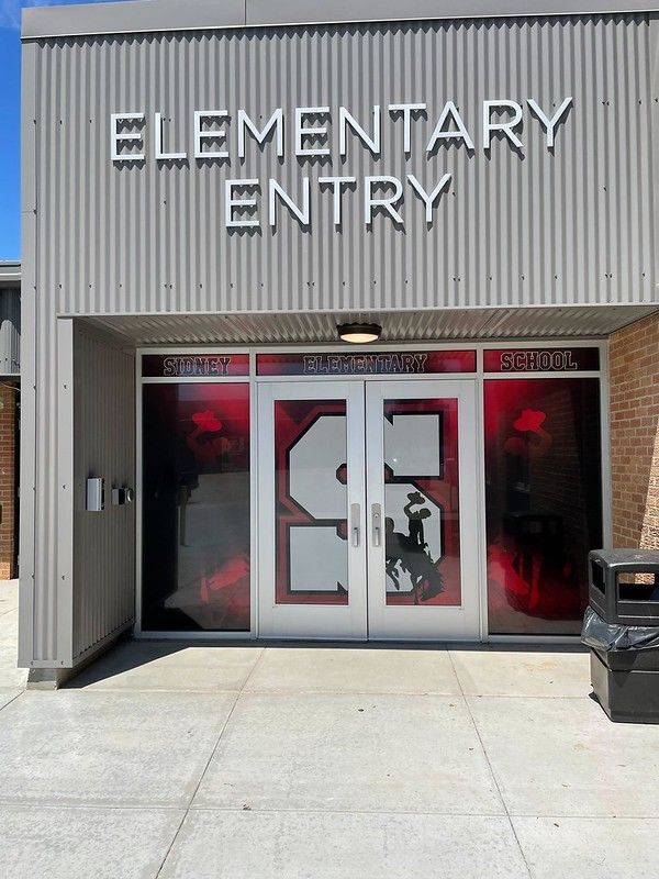 An elementary school entrance with gray metal siding and a double glass door featuring a large white letter 