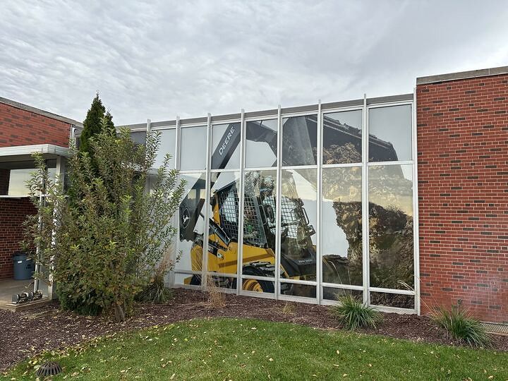 A yellow John Deere skid-steer loader reflected in the large, grid-patterned windows of a red brick building.
