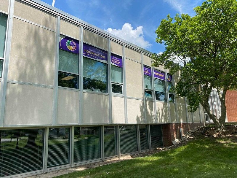 The exterior of a light-colored building with large windows and signage on a sunny day next to a leafy tree.