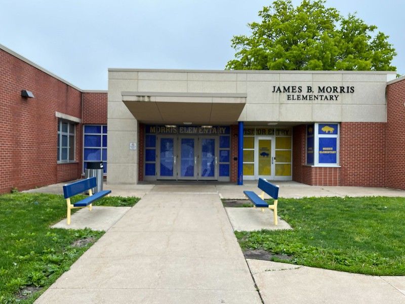 The James B. Morris Elementary school building, featuring a brick exterior, a paved walkway, and two blue benches.