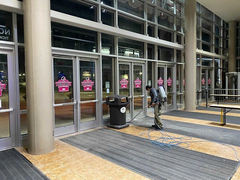 A person wearing a backpack sprayer cleans the floor in front of a building entrance with glass doors.