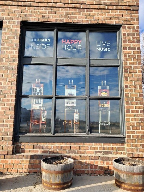 A red brick building exterior featuring a window with signs for cocktails, happy hour, live music, and liquor bottles.