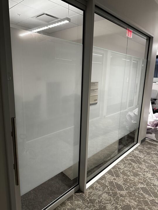 A modern office glass partition with frosted privacy film, silver door hardware, and patterned carpet flooring.