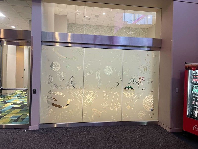 A glass wall in a corridor features frosted etchings of various geometric and scientific designs.