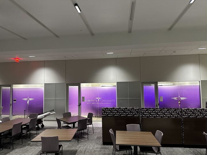 A brightly lit lounge area with several tables and chairs, featuring frosted purple glass walls with medical logos.