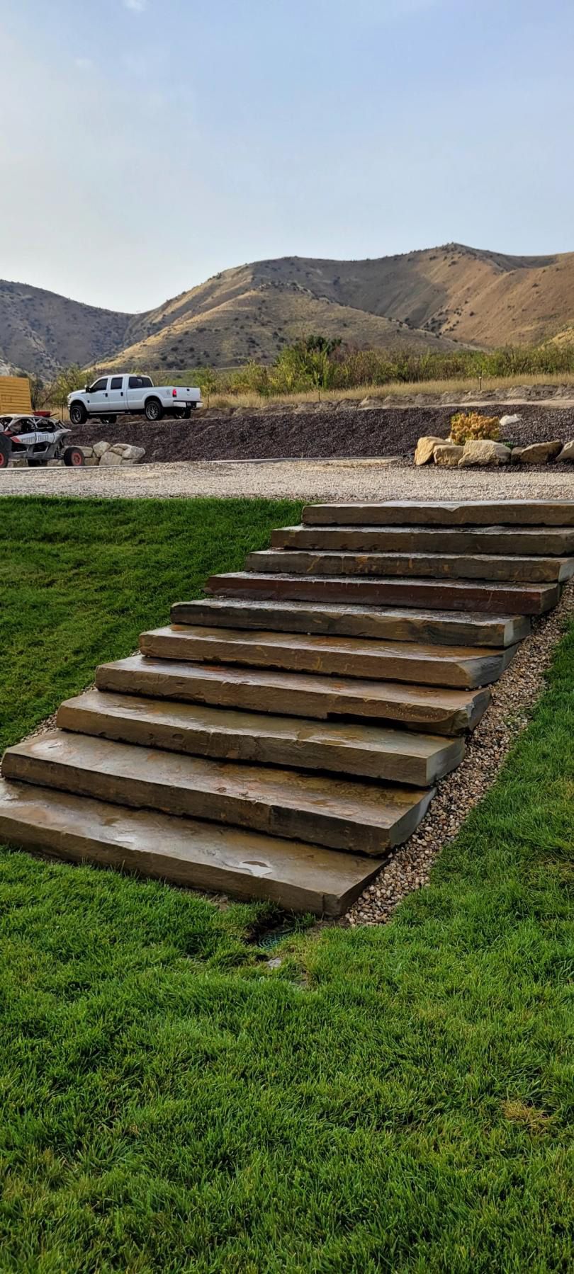 Stone steps leading through green grass toward mountains, with a white truck parked in the distance.