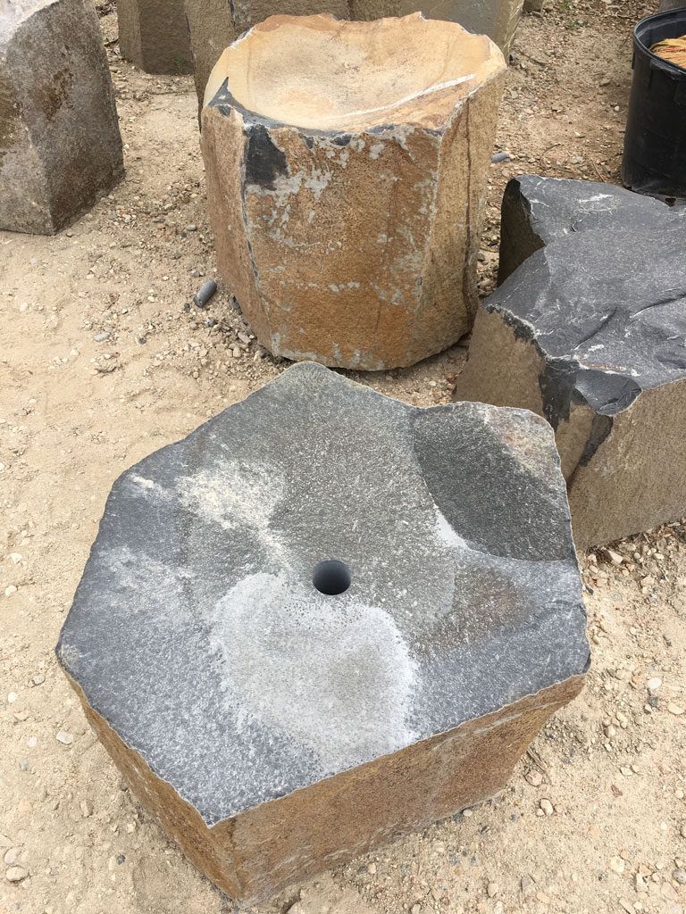 Stone blocks, some with cut sides, one with a hole, various colors, on a sandy surface.
