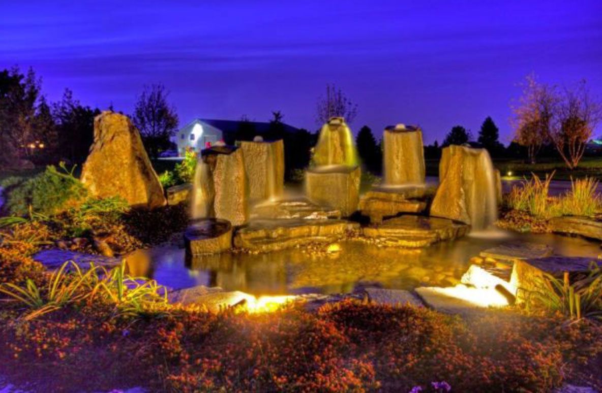 Night scene of a lit water feature with stone sculptures and reflecting pool. Blue sky.