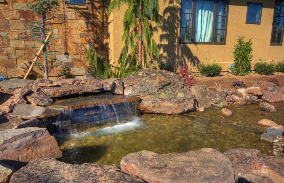 A small waterfall cascades into a pond surrounded by rocks and vegetation, next to a beige building.