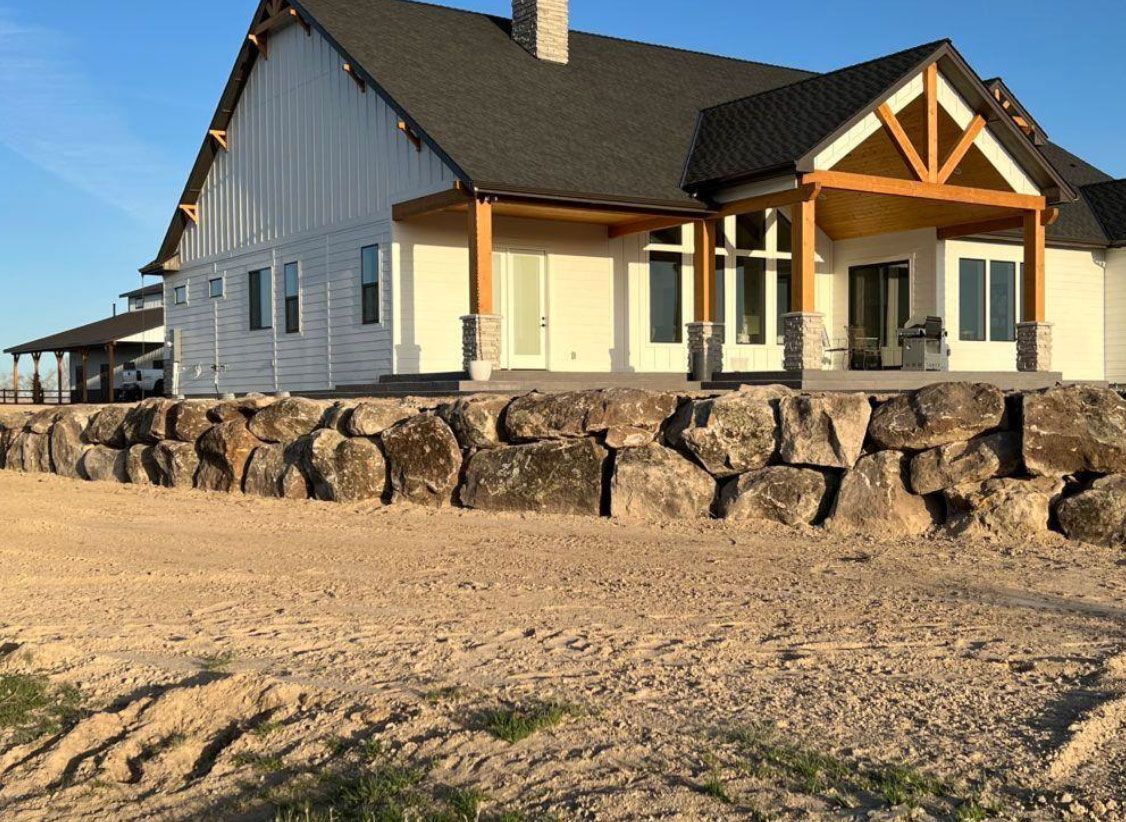 White farmhouse with wood accents, large rock wall, and a bare yard.