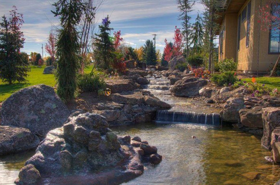 Water flows over rocks in a landscaped stream, near a house and trees.
