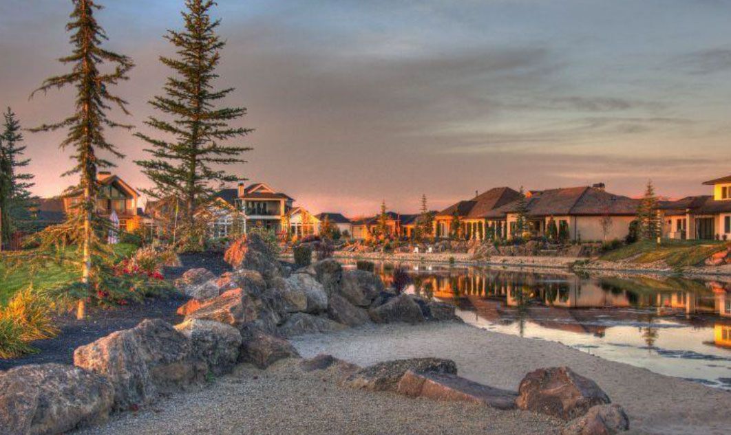 Houses line a lake at dusk, with trees and rocky shore in the foreground reflecting golden and pink light.