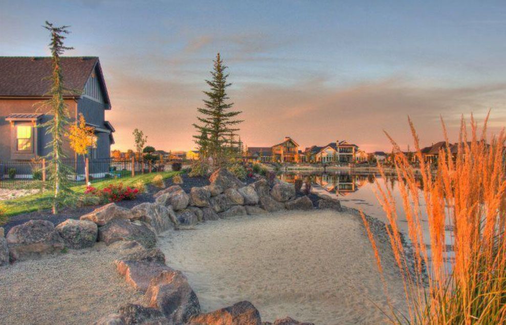 Sandy area and rocks bordering a lake, with houses in the background at sunset.
