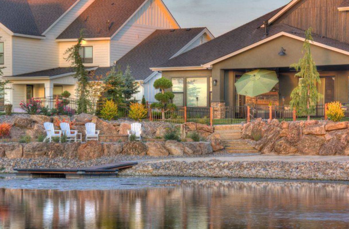 Lakeside houses with stone landscaping, white chairs, and an umbrella on the patio, reflected in calm water.
