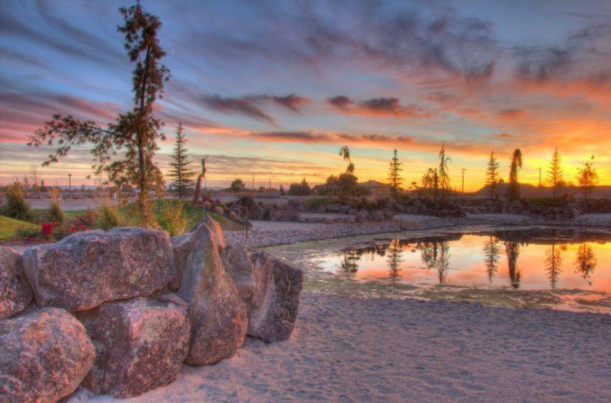 Sunset over a calm pond, reflected in the water. Large rocks in foreground, trees silhouetted in the distance.