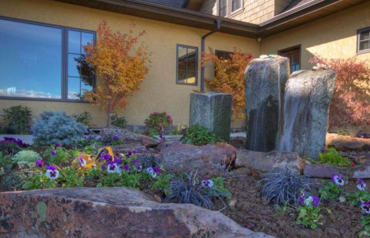Stone fountain with colorful landscaping, flowers, and autumn foliage in front of a building.