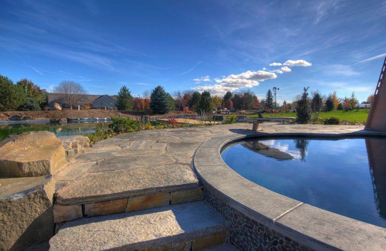 Stone patio with circular pool, water, trees, and blue sky with clouds.