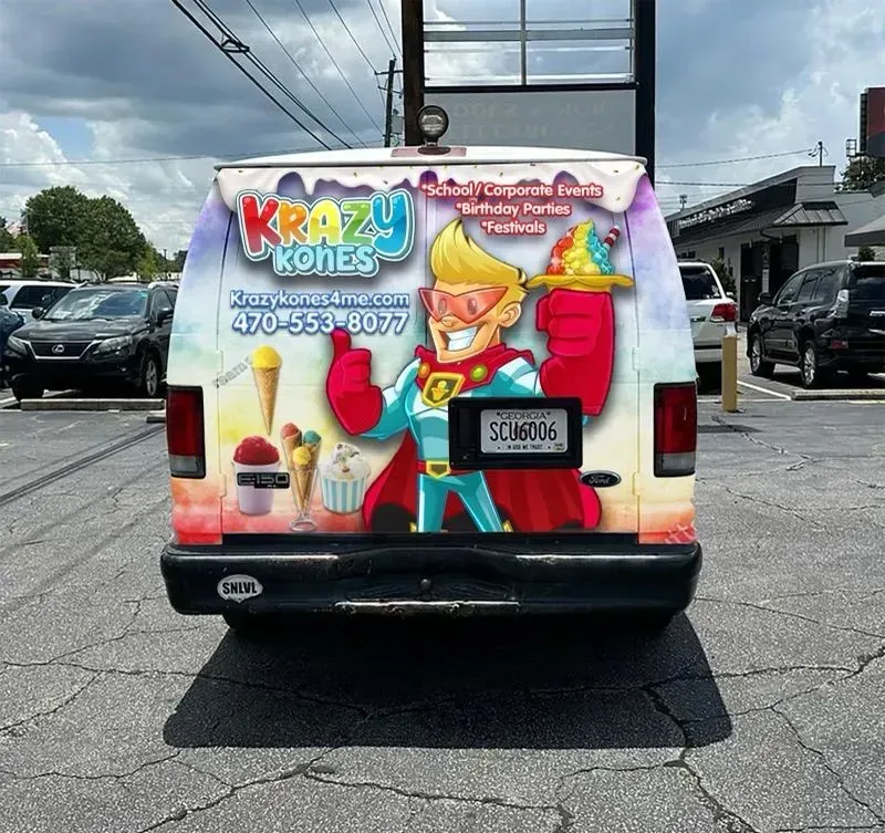 Rear view of a Krazy Kones ice cream truck with a superhero graphic. The van is in a parking lot.
