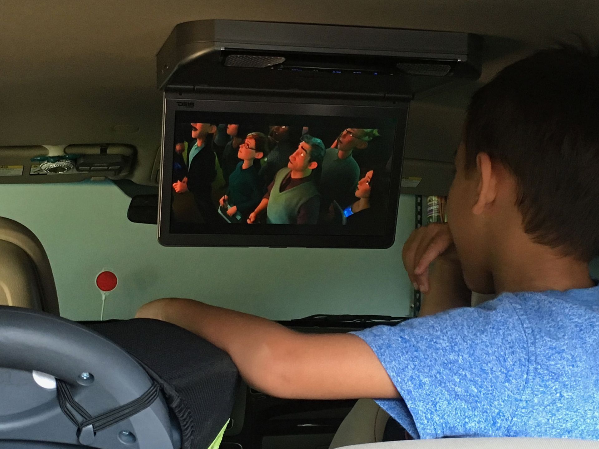 A young boy is sitting in the back seat of a car watching a movie on the overhead monitor.