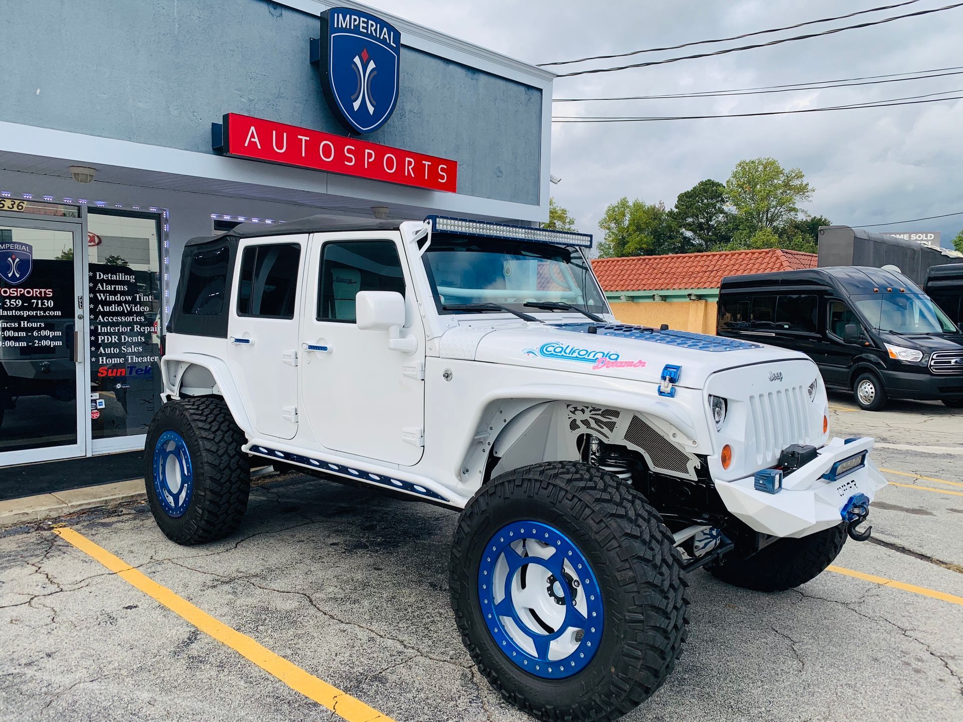 A white Jeep with blue wheels is parked in front of a car dealership.