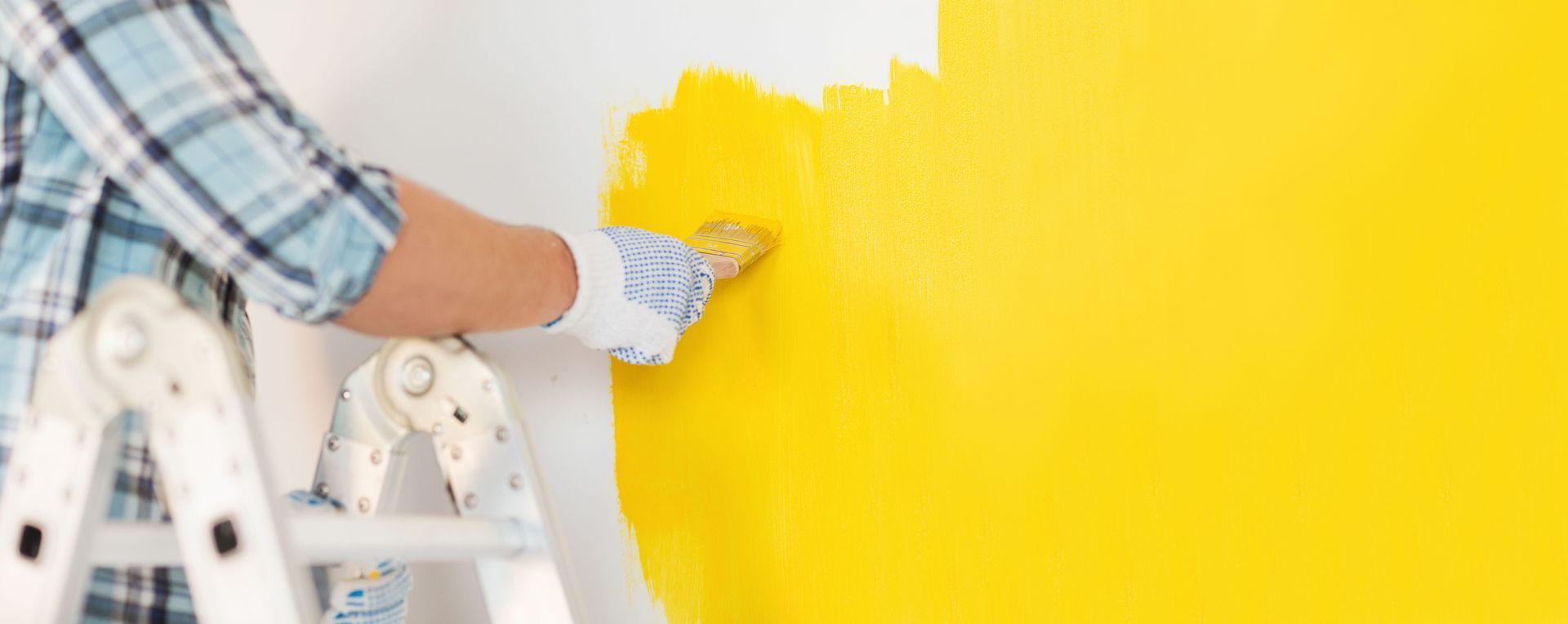 Person painting a wall yellow with a roller, standing on a step ladder.