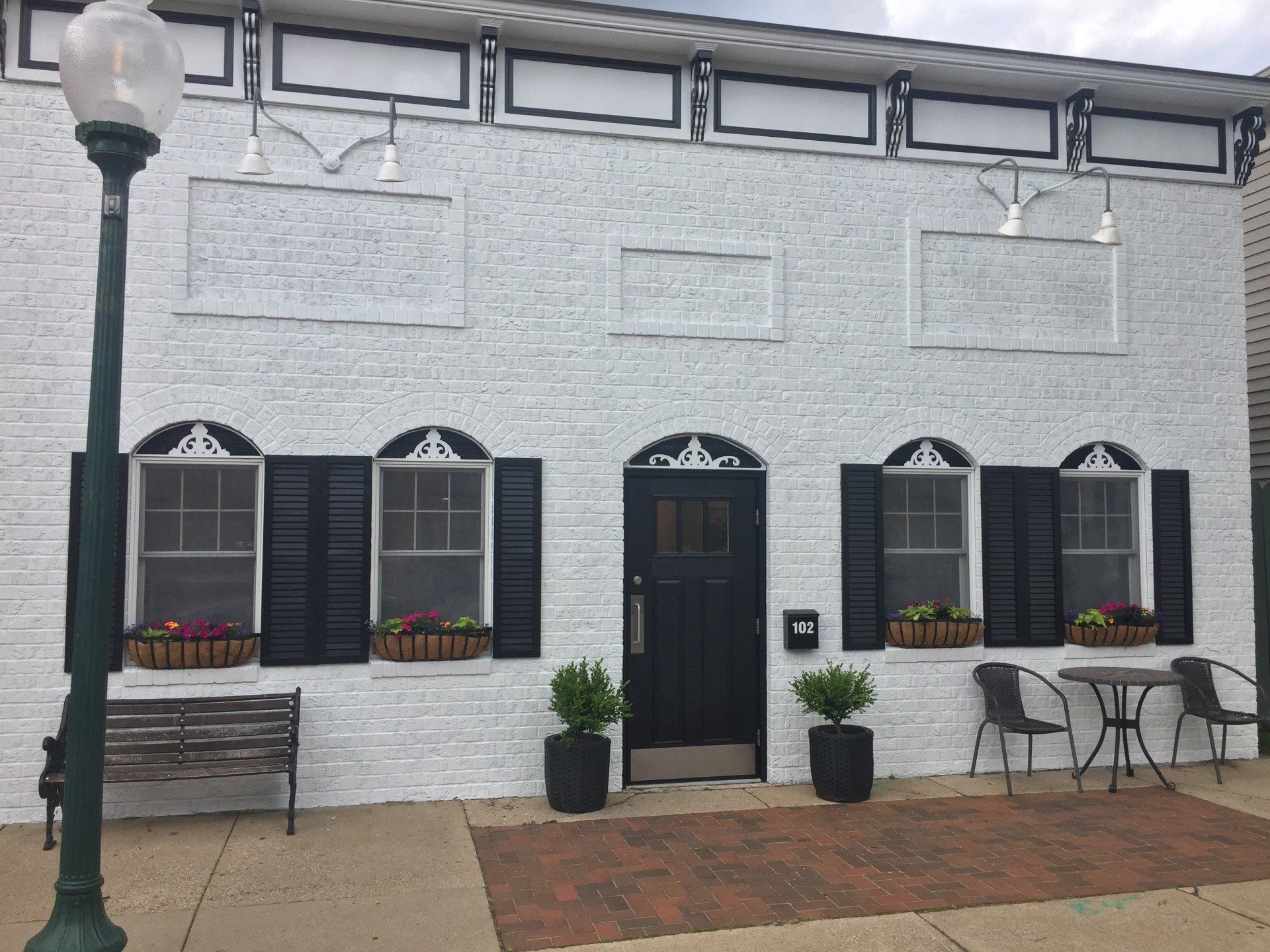 White brick building with black door and shutters, small flower boxes, and outdoor seating.