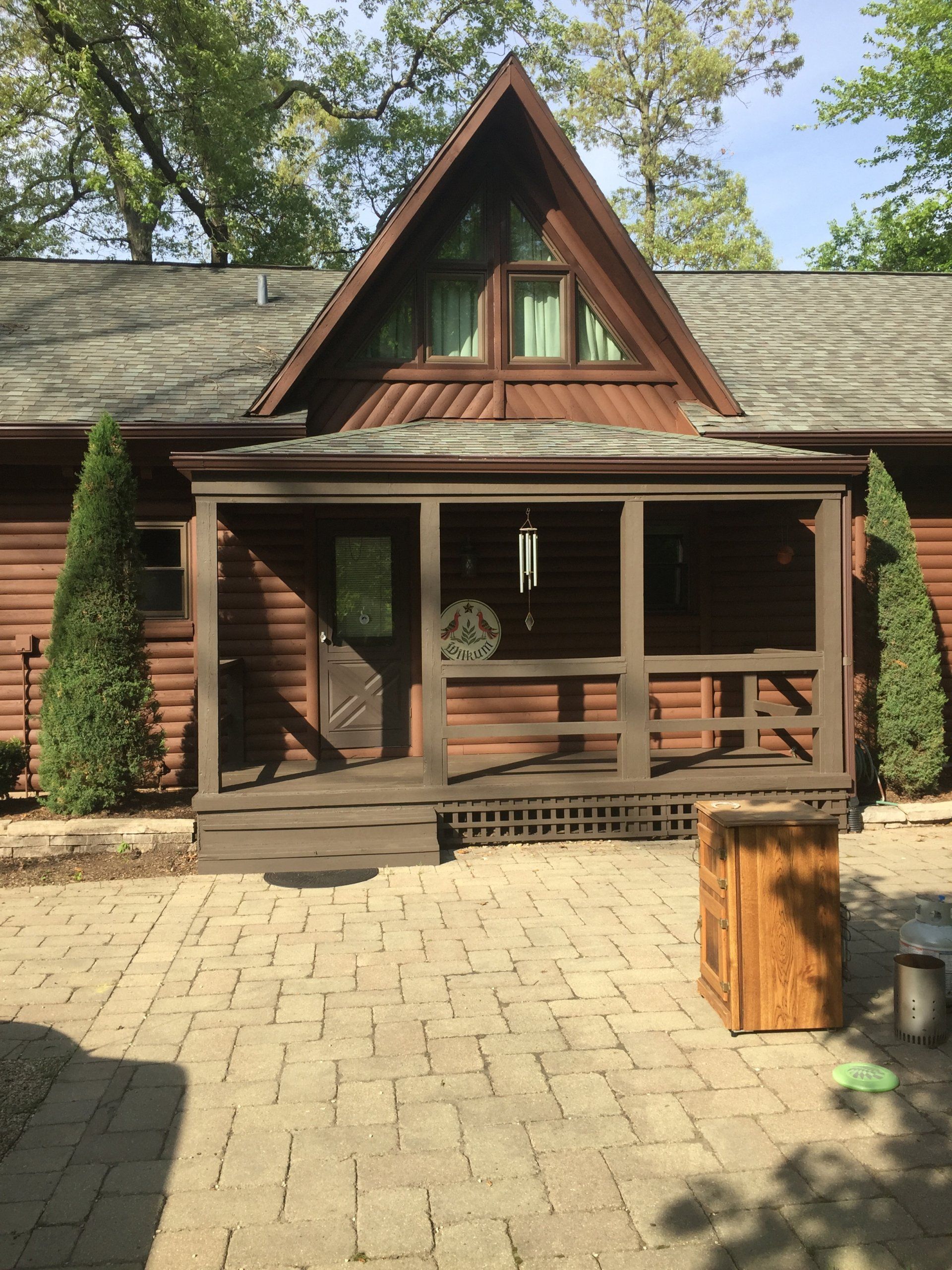 Brown log cabin with a porch and brick patio. Triangle-shaped roof above entrance.