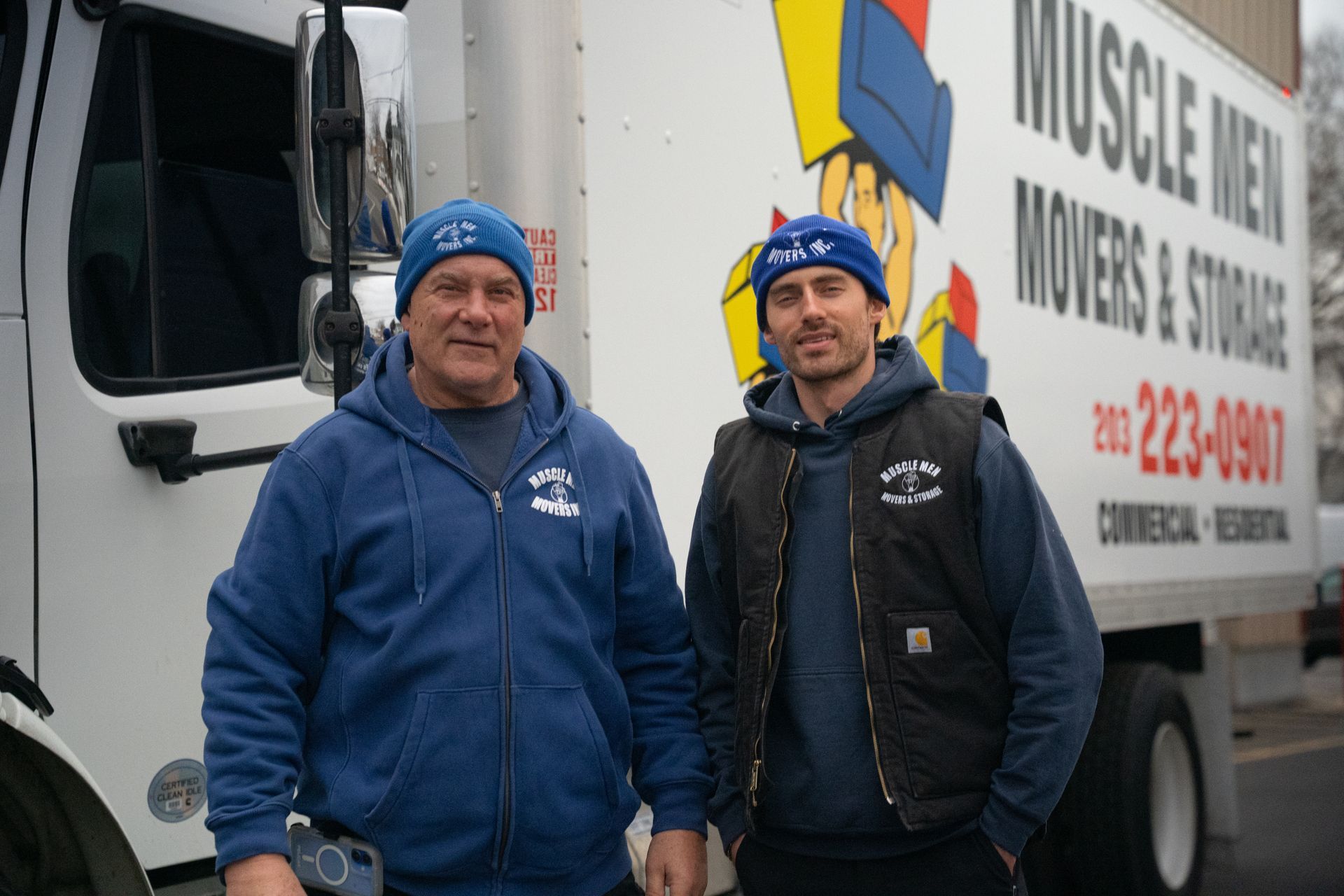 Two men in blue hats and jackets stand by a Muscle Men Movers truck.