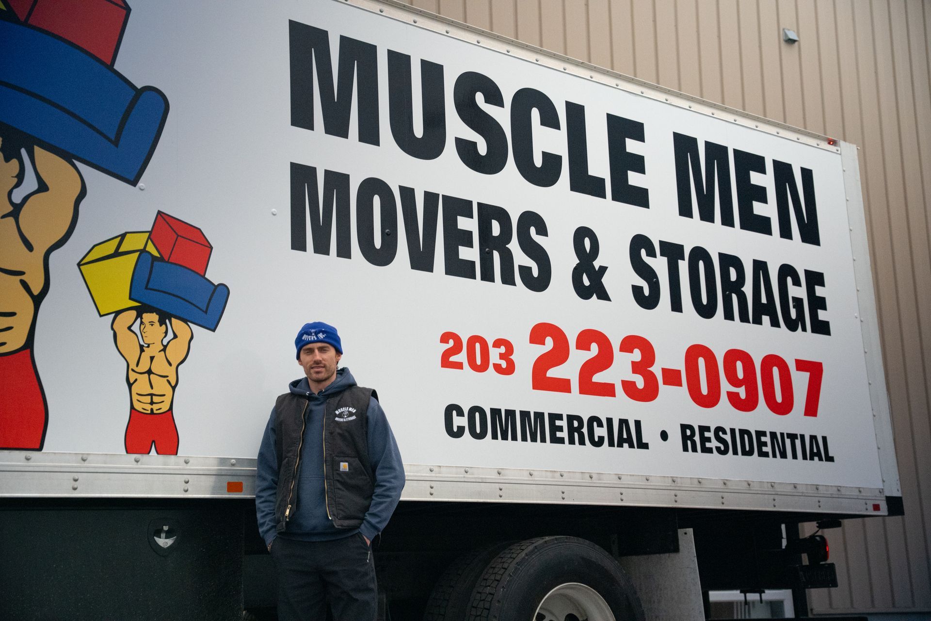 Man standing by Muscle Men Movers truck. Truck has logo and phone number.