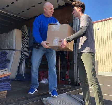 Man hands a cardboard box to another man from a truck with furniture inside.