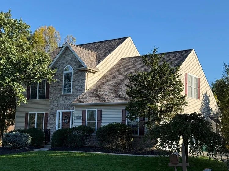 Suburban two-story house with stone facade, dark roof, and trees in front yard