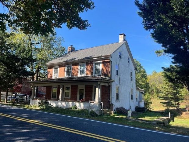 Old two-story house with white siding and red porch beside a tree-lined road on a sunny day