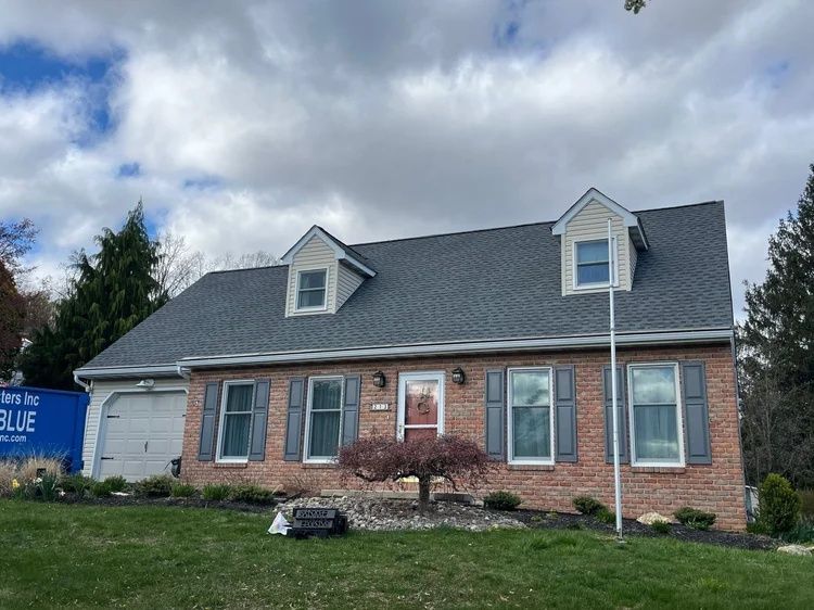 Brick house with gray roof, dormer windows, and front lawn under cloudy sky