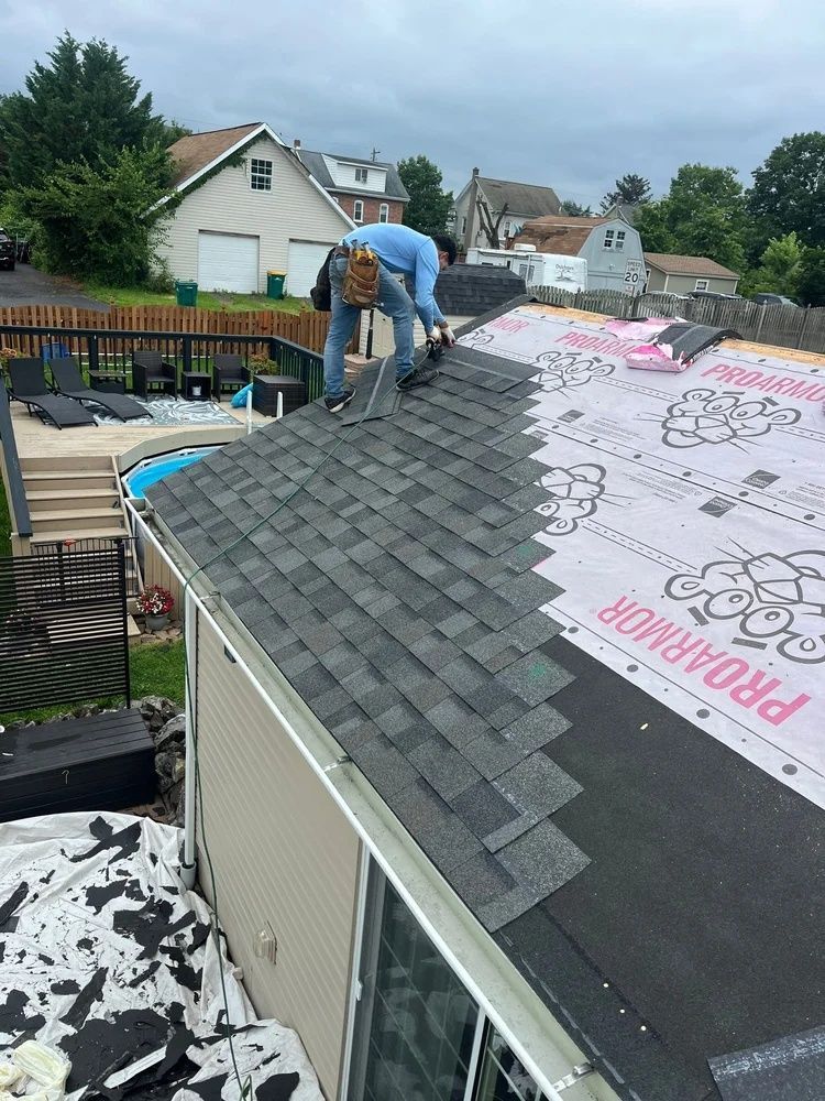 Worker installing shingles on a residential roof beside underlayment in a neighborhood.