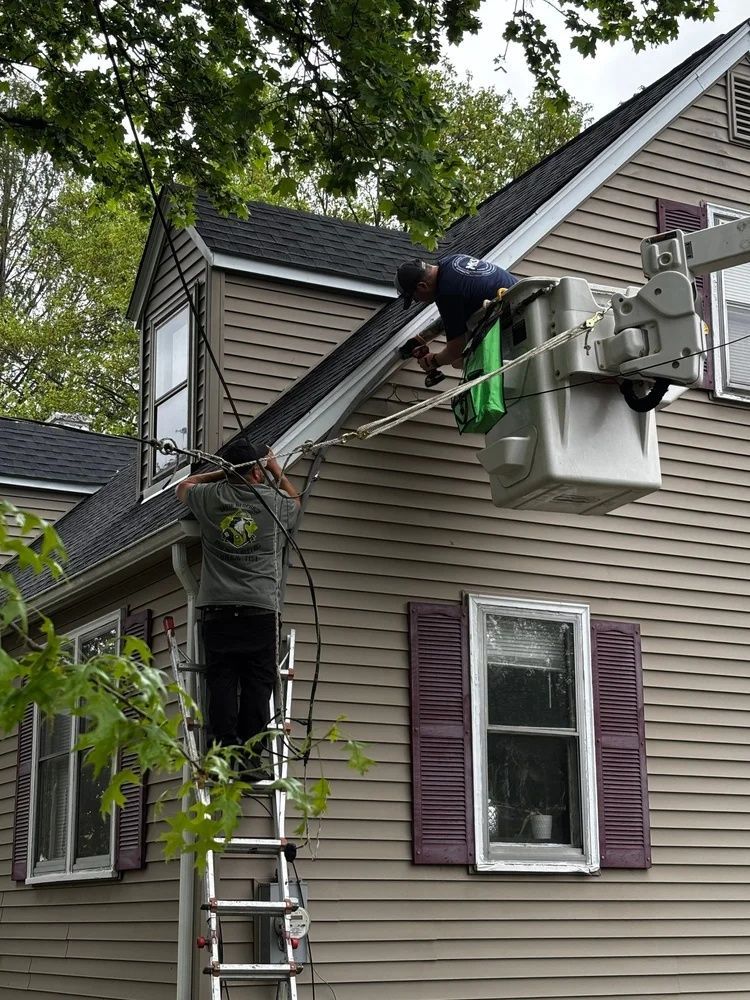 Two workers on a ladder repairing a house roof and gutter near a window with purple shutters