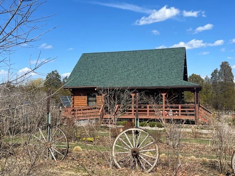 Log cabin with green roof and porch in a rural field, with an old wagon wheel in front.