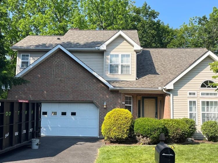 Two-story suburban house with gray siding, brick facade, white garage door, and trimmed shrubs under a blue sky