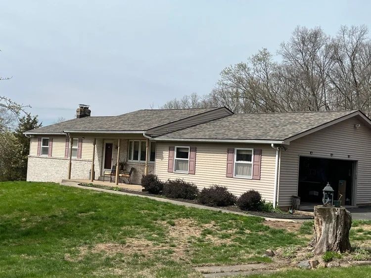 Single-story beige house with front porch, attached garage, and grassy yard under a cloudy sky
