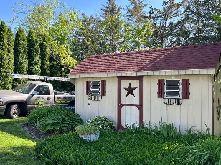 Small cream shed with red roof and star trim beside a pickup truck in a grassy yard with trees.