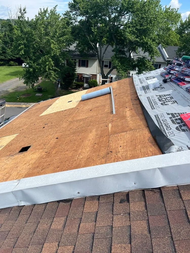 Rooftop under repair with plywood, underlayment, and shingles visible beside leafy trees and houses.