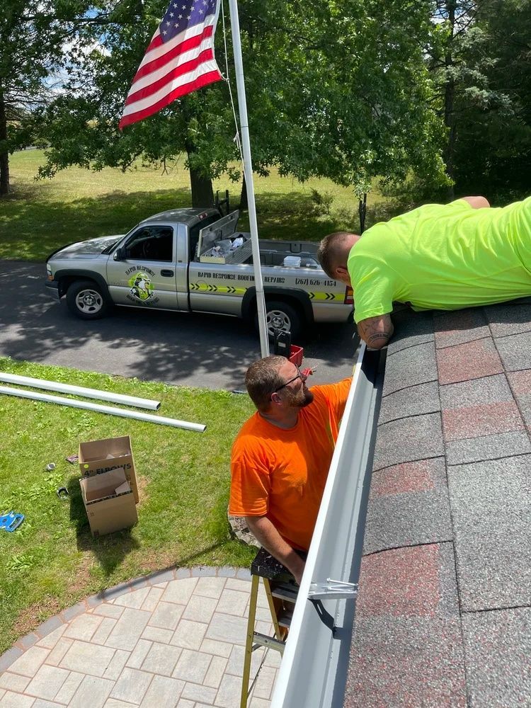 Two workers install guttering on a house roof beside a U.S. flag and parked service truck.