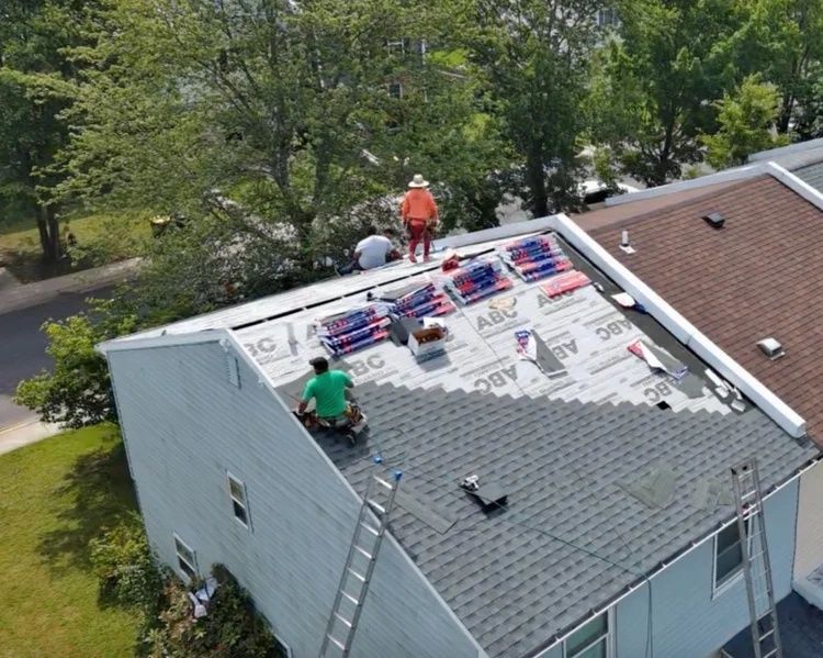 Roofers installing shingles on a house roof, with materials spread out and trees nearby.