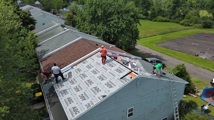 Workers installing a white roof on a house, with shingles and tools on the roof, surrounded by trees.