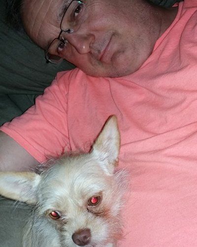 A person wearing glasses and a peach-colored shirt lying down while cuddling a small, light-colored dog.