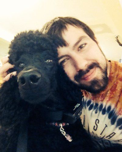 A man with dark hair and a beard leans his head against a fluffy black poodle, both looking toward the camera.