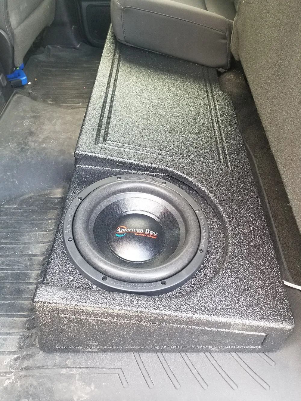 Black subwoofer installed in a vehicle, placed under the back seat.