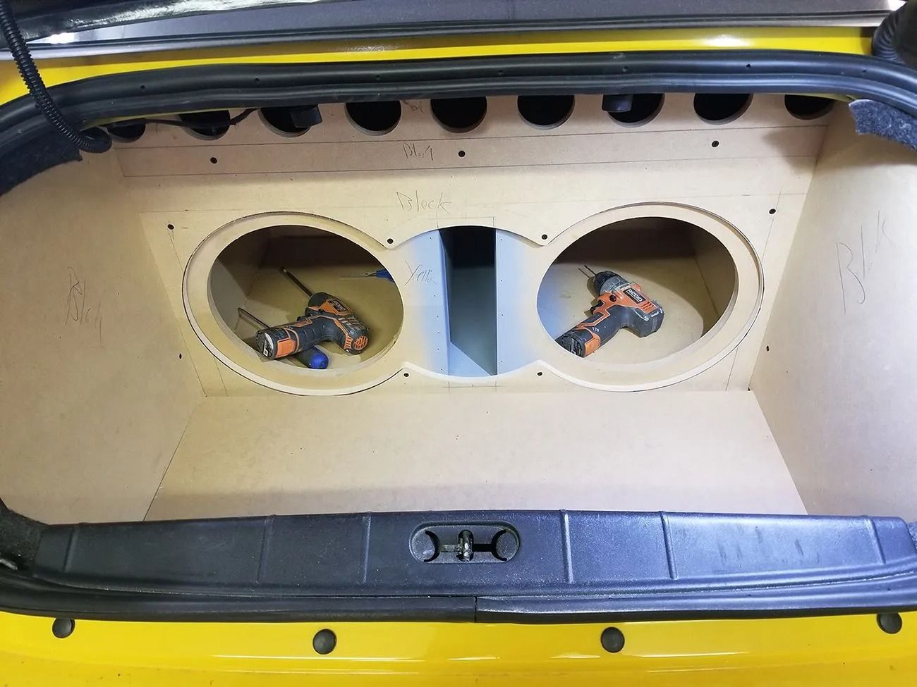Car trunk with custom wooden subwoofer enclosure, featuring two circular openings. Two drills inside.