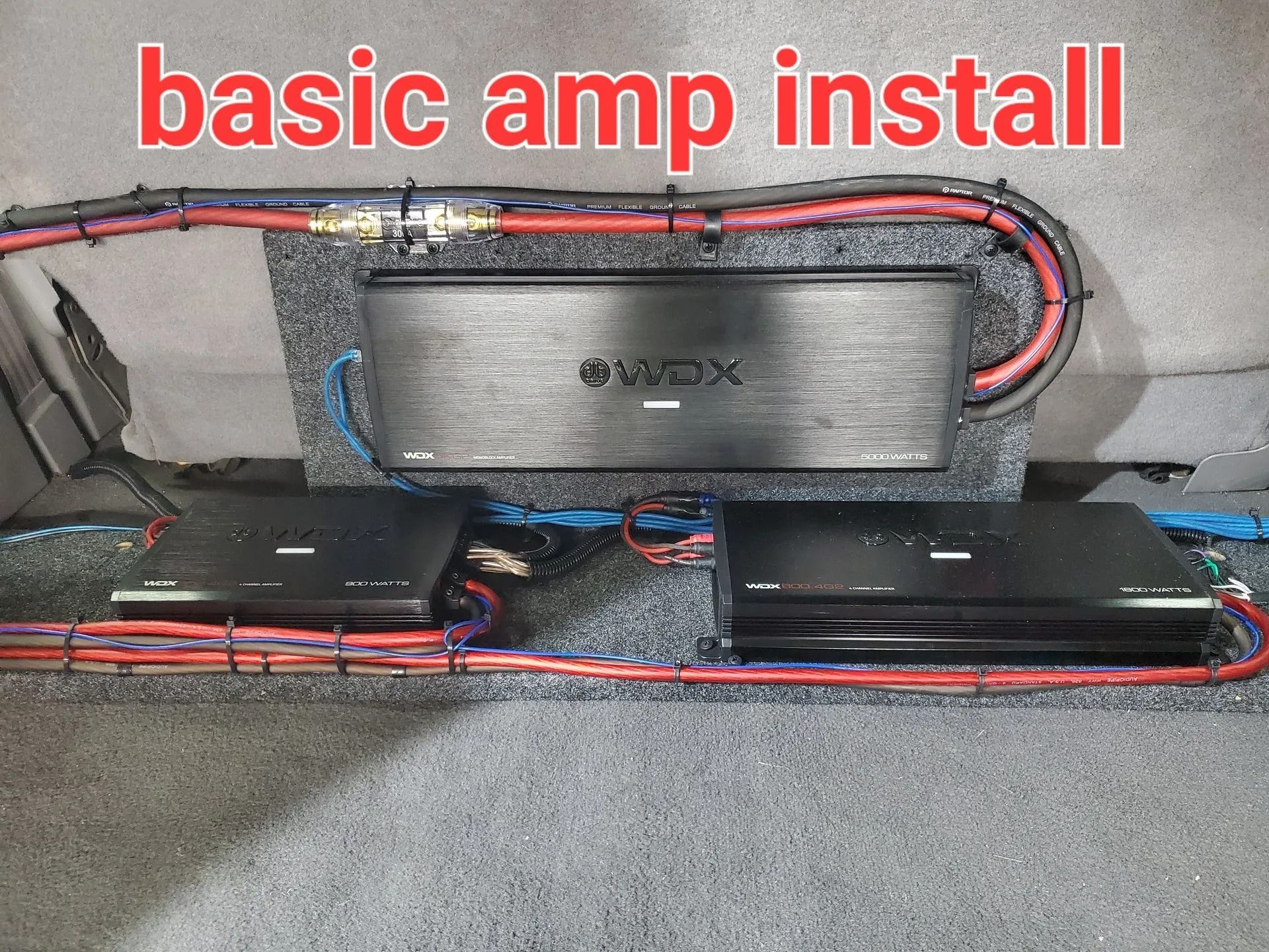 Car audio amplifier installation, red and black wiring, amplifiers mounted on a grey surface. 
